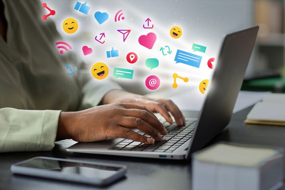 A person is typing on a laptop on a desk with floating Social media engagement Icons and emojis.